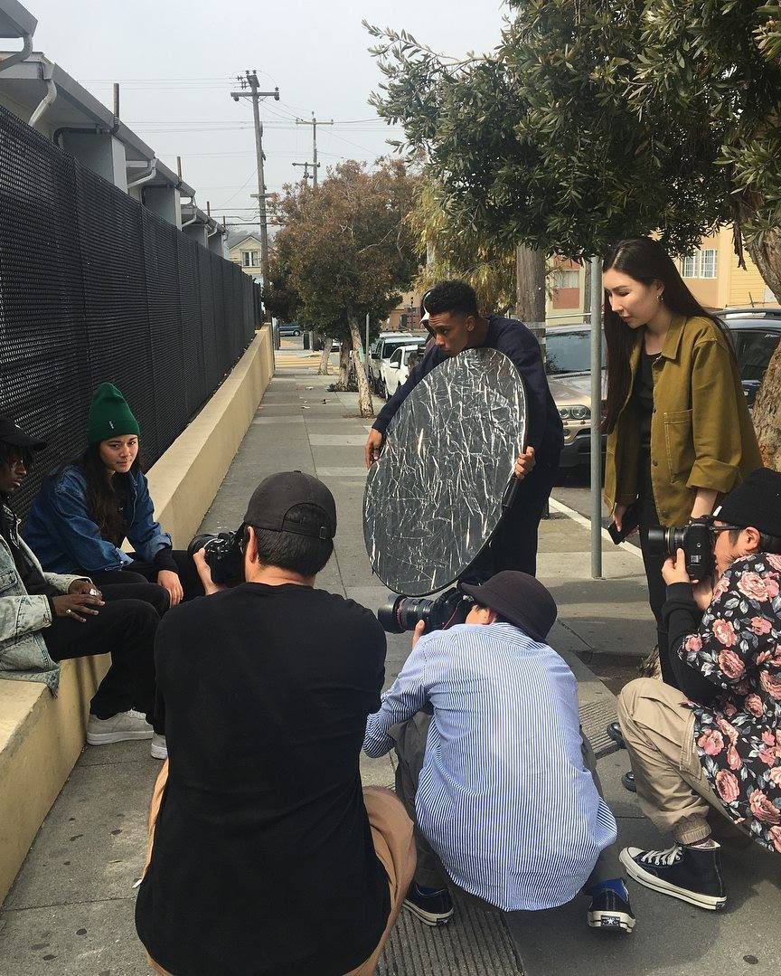 A crew gathered on a kerb on location, one member holding a circular reflector.