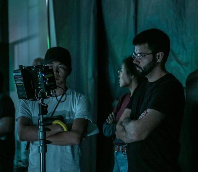 Two crew members standing behind a camera rig on a lit interior set.